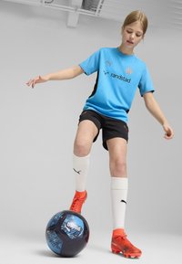 Zils īsās piedurknes krekls ar melniem akcentiem un logo, melnas bikses, baltas zeķes un spilgti oranži futbola apavi, stāv uz melnas un zilas futbola bumbas.