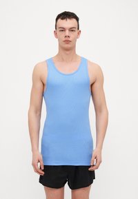 Light blue ribbed tank top with a round neckline and wide shoulder straps, paired with black shorts, model standing against a plain background.