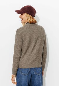 Pull en tricot marron à col haut, associé à une casquette bordeaux foncé ; porté avec un jean en denim bleu ample, mettant en avant une texture douce.