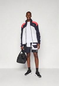 Treningsantrekk med marineblå, hvit og rød glidelåsjakke, hvit t-skjorte, svarte shorts med grafisk design og svarte joggesko. Inkluderer en svart duffelbag.