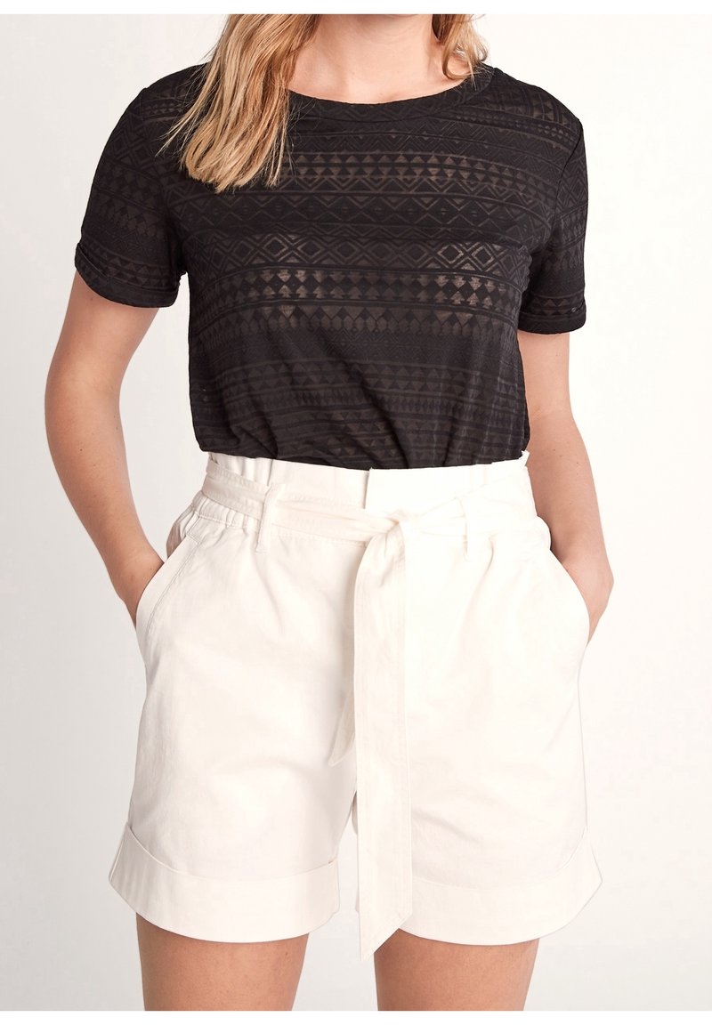 Femme portant un haut noir à manches courtes avec motif, rentré dans un short blanc taille haute ceinturé, mains dans les poches.