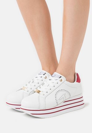 Baskets à plateforme blanches avec des rayures rouges sur la semelle et un détail rouge au talon, portées sur des jambes nues sur fond blanc.