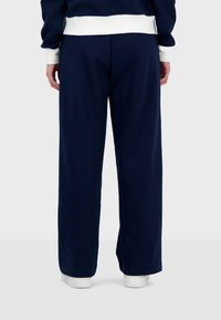Pantalones de chándal azul marino, hechos de una tela suave, con un corte relajado y un diseño de pierna recta, combinados con zapatillas blancas sobre un fondo neutro.
