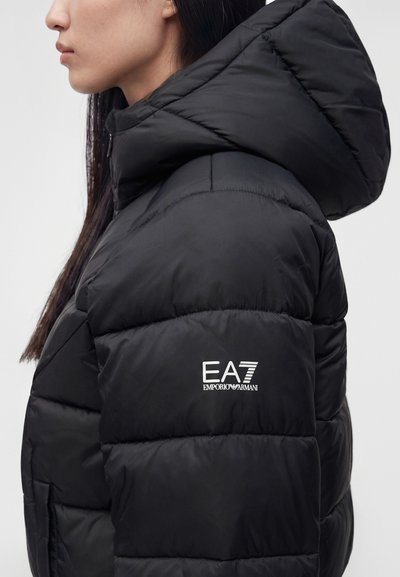 Страничен профил на човек, носещ черно пухено яке с качулка и бяло лого EA7 Emporio Armani на горната част на ръкава.