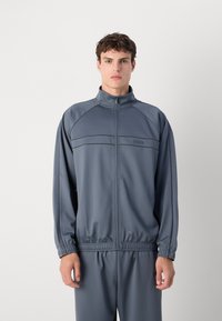Lett grå jakke til trening med høy krage, glidelås og kontrasterende sømmer. Har en fitted midje og matchende bukser.