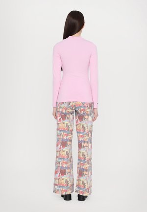 Femme aux longs cheveux foncés portant un haut rose à manches longues et un pantalon imprimé collage coloré, debout de dos sur un fond blanc uni.