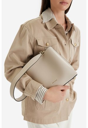 Frau trägt eine beige Jacke über einem gestreiften Hemd und hält eine hellbeige Gino Rossi Clutch mit goldenen Buchstaben und Trageriemen.