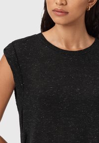 Donna che indossa un top senza maniche nero a pois con scollatura rotonda e piccoli orecchini ad anello dorati, su uno sfondo chiaro e uniforme.