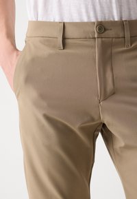 Pantaloni bej cu o textură fină, având închidere cu nasture, bucle pentru curea și buzunare laterale. Țesătura pare ușoară și elegantă.