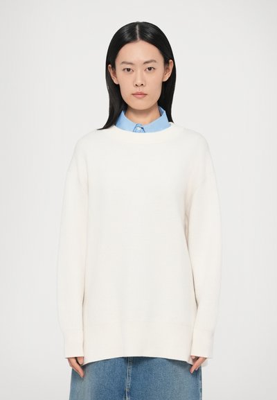 Pull blanc oversize avec un col rond et des épaules tombantes, associé à une chemise à col bleu clair. Texture lisse en maille.