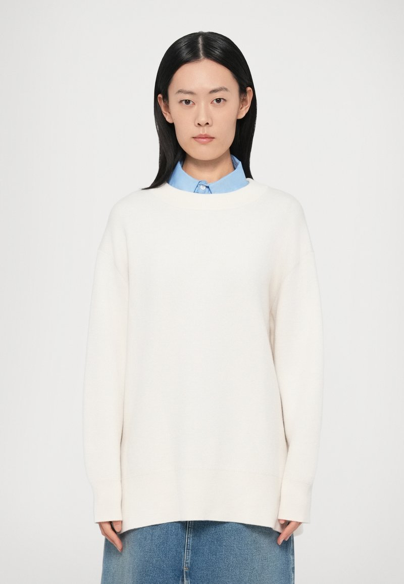 Fehér, túlméretezett pulóver kerek nyakkivágással és ejtett vállakkal, világoskék galléros inggel párosítva. Sima, kötött text�úra.