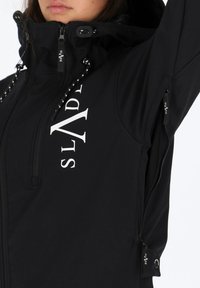 Czarna kurtka z kapturem z białym pionowym logo "SLYDE", z kieszeniami na zamek pod ramieniem i na klatce piersiowej, noszona przez osobę z uniesioną ręką.