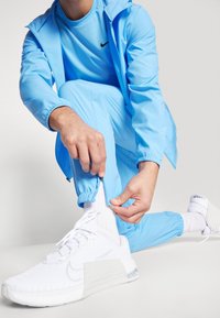 Lichtblauwe sporttracksuit met een ritssluiting bij de manchetten, gecombineerd met witte sneakers met gestructureerde stof en een grijze accenten.
