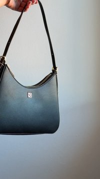 Hand hält eine schwarze Hugo Schultertasche aus Leder mit goldenem Reißverschluss und Beschlägen vor einem schlichten, hellen Hintergrund.