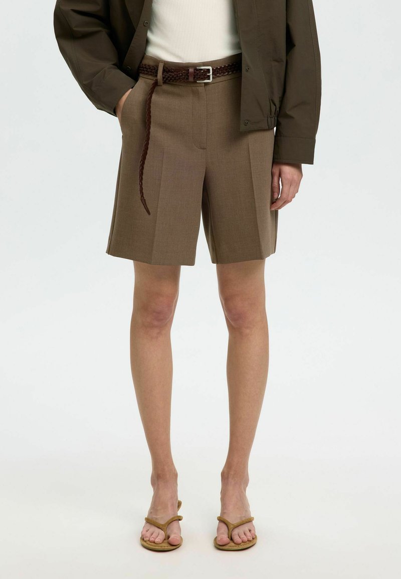Femme portant un short marron sur mesure longueur genou avec une ceinture tressée, une veste olive, un haut blanc et des sandales marron, debout devant un fond blanc.