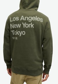 Olivengrøn hættetrøje med store hvide bogstaver: "Los Angeles," "New York," og "Tokyo" på ryggen. Blødt bomuldsstof med en afslappet pasform.