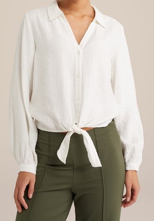 Camisa blanca de manga larga con botones, anudada a la cintura, combinada con pantalones de tiro alto de color verde oliva, mostrando solo el torso y los brazos.