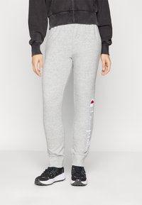 Grå bomullsjoggebukser med elastisk midje, innsnevring ved anklene, og en hvit brodert logo langs siden. Kombinert med svarte joggesko.