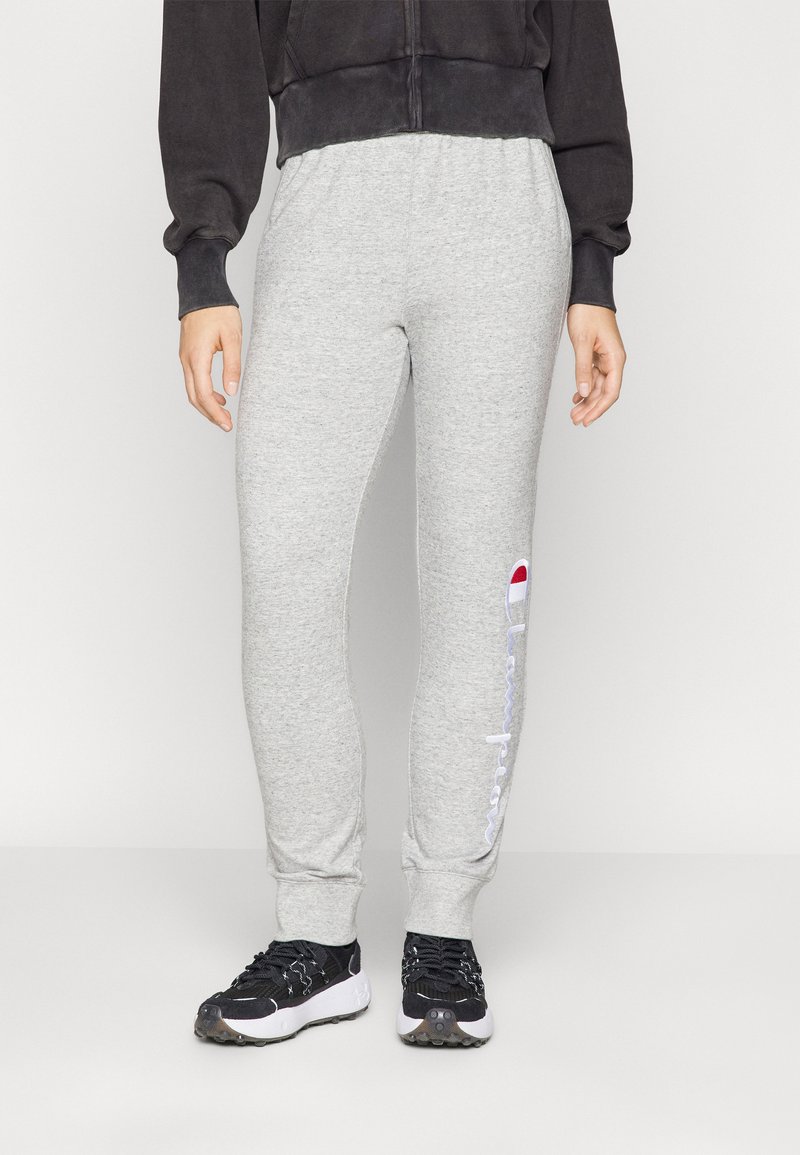 Grå bomullsjoggebukser med elastisk midje, innsnevring ved anklene, og en hvit brodert logo langs siden. Kombinert med svarte joggesko.