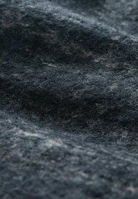 Tmavě šedá vlněná tkanina s jemnou, načechranou strukturou a drobnými volnými vlákny, zobrazená v detailu s jemnými záhyby a stíny.