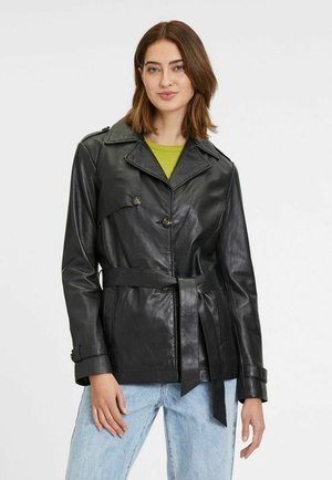 Zwarte leren jas met een ceintuur in de taille, dubbelzijdig ontwerp, grote kraag en voorzakken, gedragen over een groene top en lichte jeans.