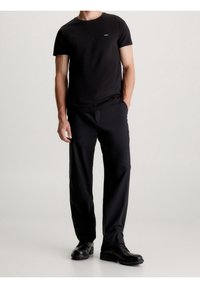 Musta pyöreäkauluksinen t-paita, jossa pieni logo, yhdistettynä löysästi istuviin mustiin housuihin ja mustiin nauhakenkäisiin saappaisiin, luoden yksivärisen ilmeen.