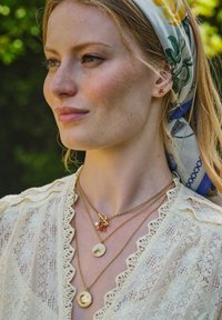 Woman with light freckles wearing a floral headscarf, cream lace blouse, and layered gold necklaces with round pendants and a rose charm.