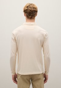Helles beige Poloshirt mit langen Ärmeln und Kragen, mit einer glatten Textur und einem kleinen rechteckigen Akzent auf der Rückseite.