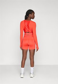 Žena s pletenicom u kosi, obučena u svijetlu narančastu Nike Pro dugu majicu i šorts, bijele čarape i bijele tenisice, stoji okrenuta leđima.