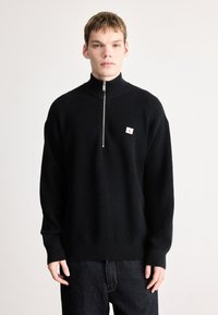 Čierny pletený pulóver s vysokým golierom a predným zipsom, s malou bielo-červenou logovou nášivkou na hrudi. Tmavé džínsové nohavice.