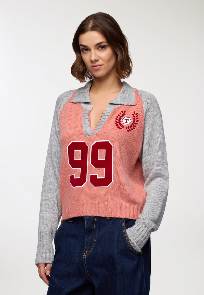 Giovane donna che indossa un maglione a maglia rosa e grigio con "99" rosso sul davanti e jeans blu scuro, in posa con una mano in tasca.