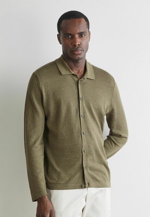 Mann trägt olivgrünen, geknöpften Cardigan über weißen Hosen, steht vor schlichtem hellem Hintergrund, mit einer Hand hinter dem Rücken.