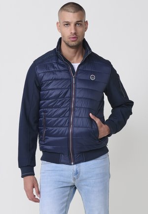 Jachetă puffer navy cu fermoar frontal, textură matlasată și buzunare laterale. Dispune de mâneci tricotate și un logo rotund pe piept.