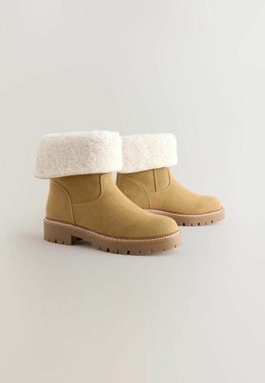Sandfarbene Wildleder-Ankle-Boots mit einem cremefarbenen Kunstfellumschlag, strukturiertem Sohlenprofil und kontrastierendem Stitching um die Sohle. Abgerundetes Zehen-Design.