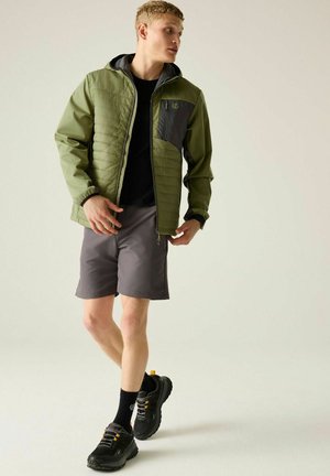 Young man wearing a green jacket, black shirt, gray shorts, black socks, and black sneakers, looking to the side while walking.