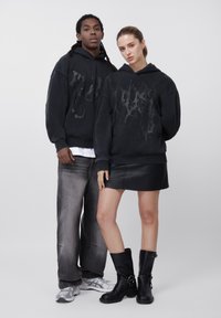 Dos modelos con sudaderas con capucha negras con estampados gráficos; un hombre con vaqueros desgastados y zapatillas, una mujer con falda de cuero negra y botas negras sobre fondo gris.