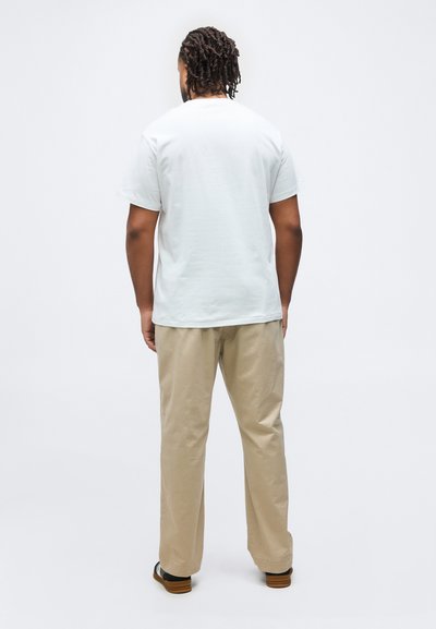 Άνδρας με κοτσίδες στέκεται στραμμένος προς τα πίσω φορώντας ένα απλό λευκό μπλουζάκι, μπεζ παντελόνι και μαύρα και λευκά παπούτσια σε λευκό φόντο.