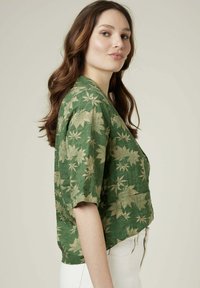 Blouse verte à manches courtes avec un motif de feuilles, fabriquée en tissu texturé. Présente une coupe décontractée et un décolleté en V profond.