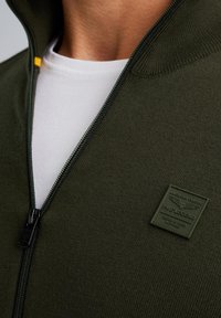 Dunkelgrüner Reißverschluss-Pullover mit geripptem Kragen und strukturiertem Finish. Verfügt über einen schwarzen Reißverschluss und ein erhabenes Logo-Patch auf der Brust.