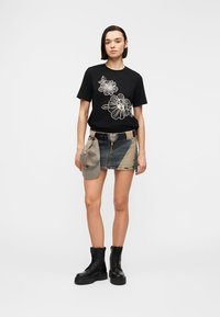 Femme portant un t-shirt noir à motifs floraux, une mini-jupe en jean patchwork avec ceinture à boucle en forme de cœur, tenant une casquette grise et des bottes de combat noires sur fond blanc.
