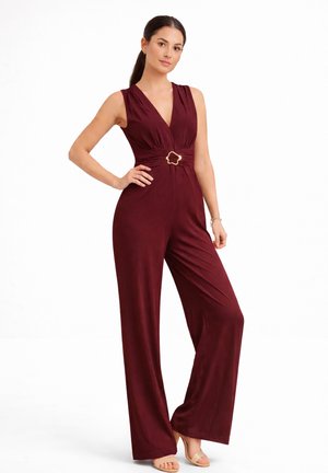 Kobieta stoi, ubrana w bezrękawny bordowy kombinezon z szerokimi nogawkami i ozdobną złotą klamrą w talii, w parze z beżowymi szpilkami.
