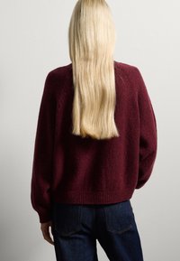 Suéter de punto burdeos con un corte holgado, dobladillo acanalado y mangas largas, combinado con jeans de mezclilla azul oscuro. Textura suave y tejido blando.
