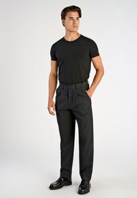 Svart åtsittande t-shirt i kombination med högmidjade, mörkgrå byxor med veck. Modellen bär blanka svarta skor. Enkelt och modernt design.