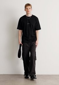 Joven vestido con una camiseta negra de Hugo, pantalones cargo oscuros, zapatos negros y sosteniendo una pequeña bolsa negra, de pie sobre un fondo sencillo.