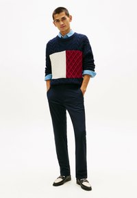 Maglione blu navy a maglia grossa con un motivo geometrico testurizzato in bordeaux e bianco; abbinato a pantaloni scuri e mocassini neri con dettagli bianchi.