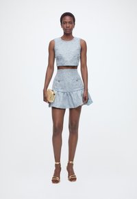 Γυναίκα που φοράει ανοιχτό μπλε αμάνικο κοντό τοπ και ταιριαστή φούστα με καμπάνα, κρατώντας ένα χρυσό τσαντάκι και φορώντας χρυσά σανδάλια με λουράκια.