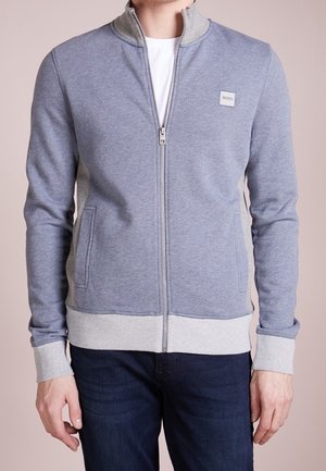 Uomo che indossa una giacca leggera grigio chiaro con cerniera e tasche laterali, con un piccolo logo BOSS sul petto, abbinata a jeans blu scuro.