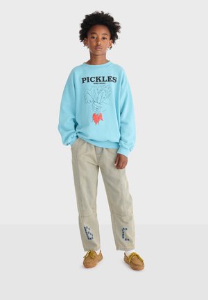 Bambino con capelli ricci indossa una felpa blu con la scritta "PICKLES", pantaloni beige con motivi blu sulle ginocchia e mocassini marroni con lacci gialli.