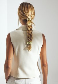 Sleeveless cream blazer with a fitted design. Hair in a braid secured by a gold hair accessory. Visible tattoo on the left arm.
