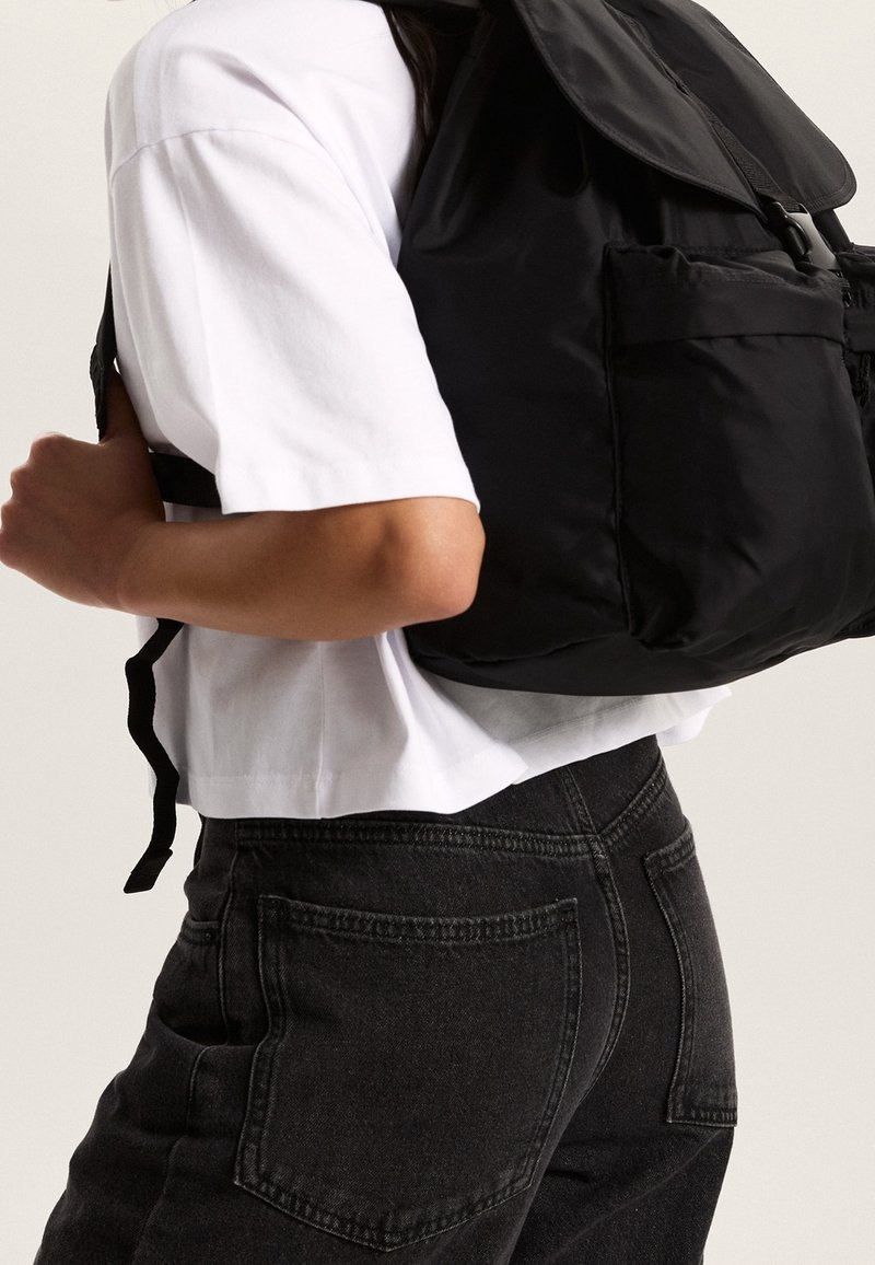 Personne portant une chemise blanche à manches courtes et un jean noir, portant un grand sac à dos noir avec des poches frontales dans le dos.